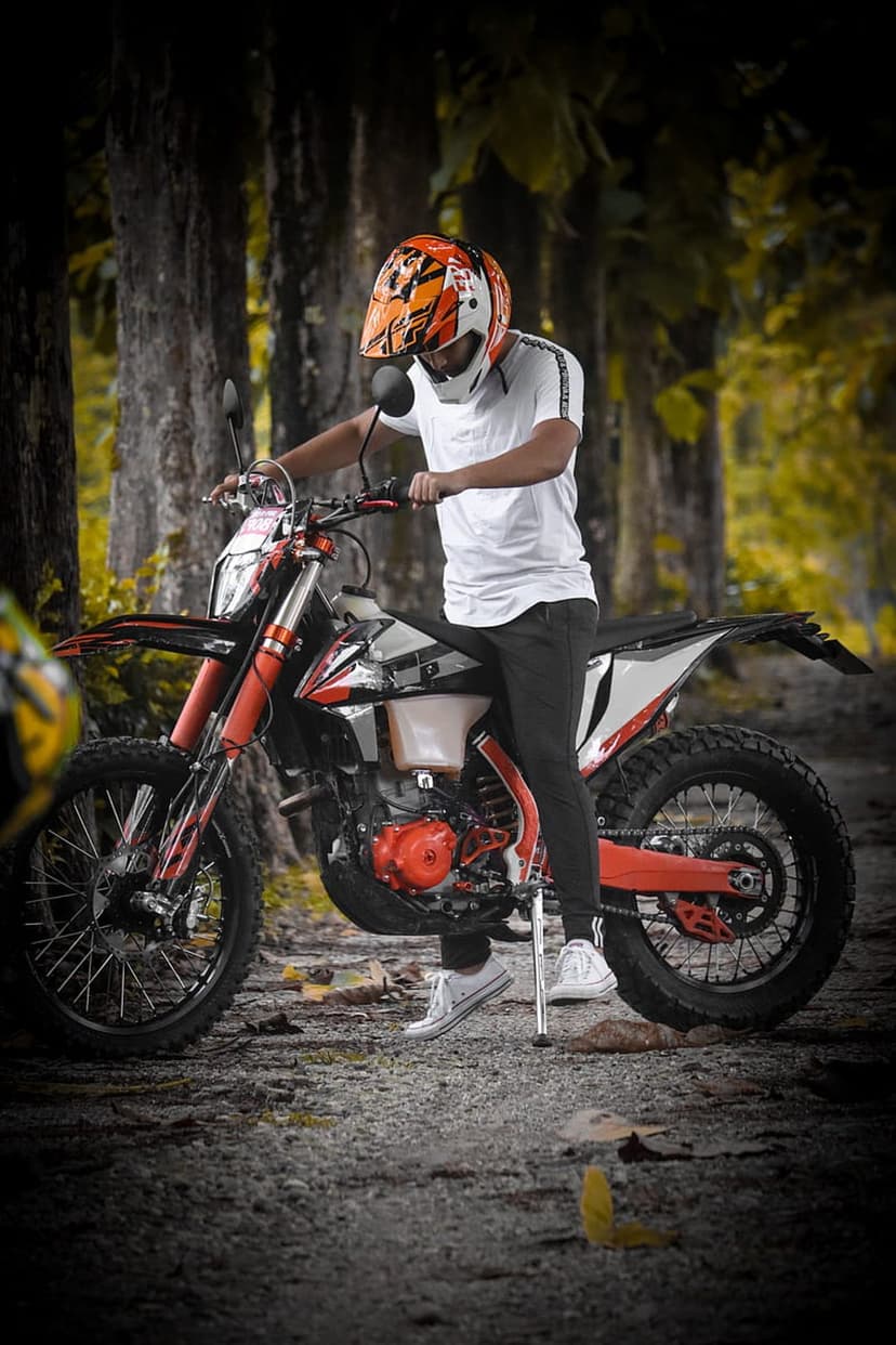 Motocross rider in an orange helmet sits on a red dirt bike along a wooded trail with autumn foliage.