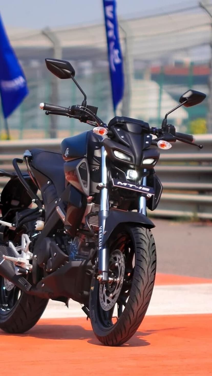 Yamaha MT-15 black motorcycle stands on an orange racetrack under bright daylight with blue flags behind it.