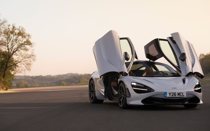 Silver McLaren 720S supercar with open dihedral doors on a race track at sunset under a soft golden sky.