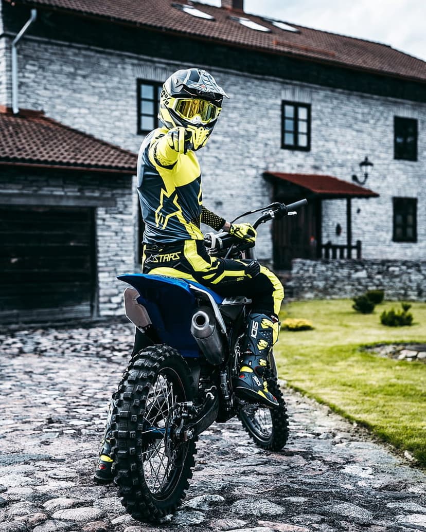 Motocross rider in yellow and black gear sits on a dirt bike over cobblestones near a rustic stone building.