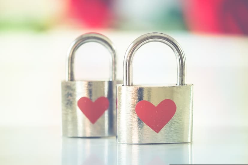 Twin silver padlocks with small red heart stickers resting against a soft pink and green bokeh backdrop.