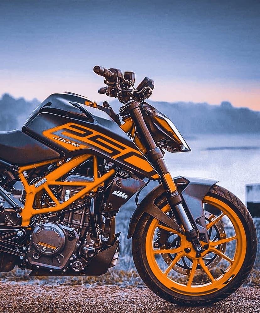 KTM 250 Duke motorcycle with orange wheels and black tank parked outdoors against a blurred water background.