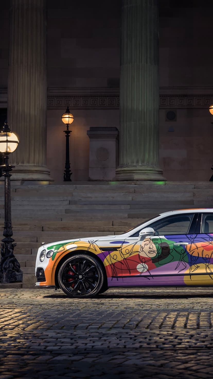 Bentley Continental GT with colorful abstract livery parked on a dark cobblestone street under yellow lamps.