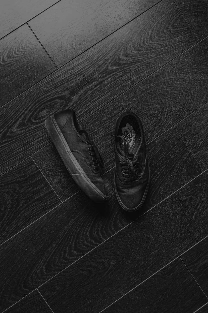 Classic canvas sneakers rest on dark diagonal wood floorboards in a high-angle black and white composition.