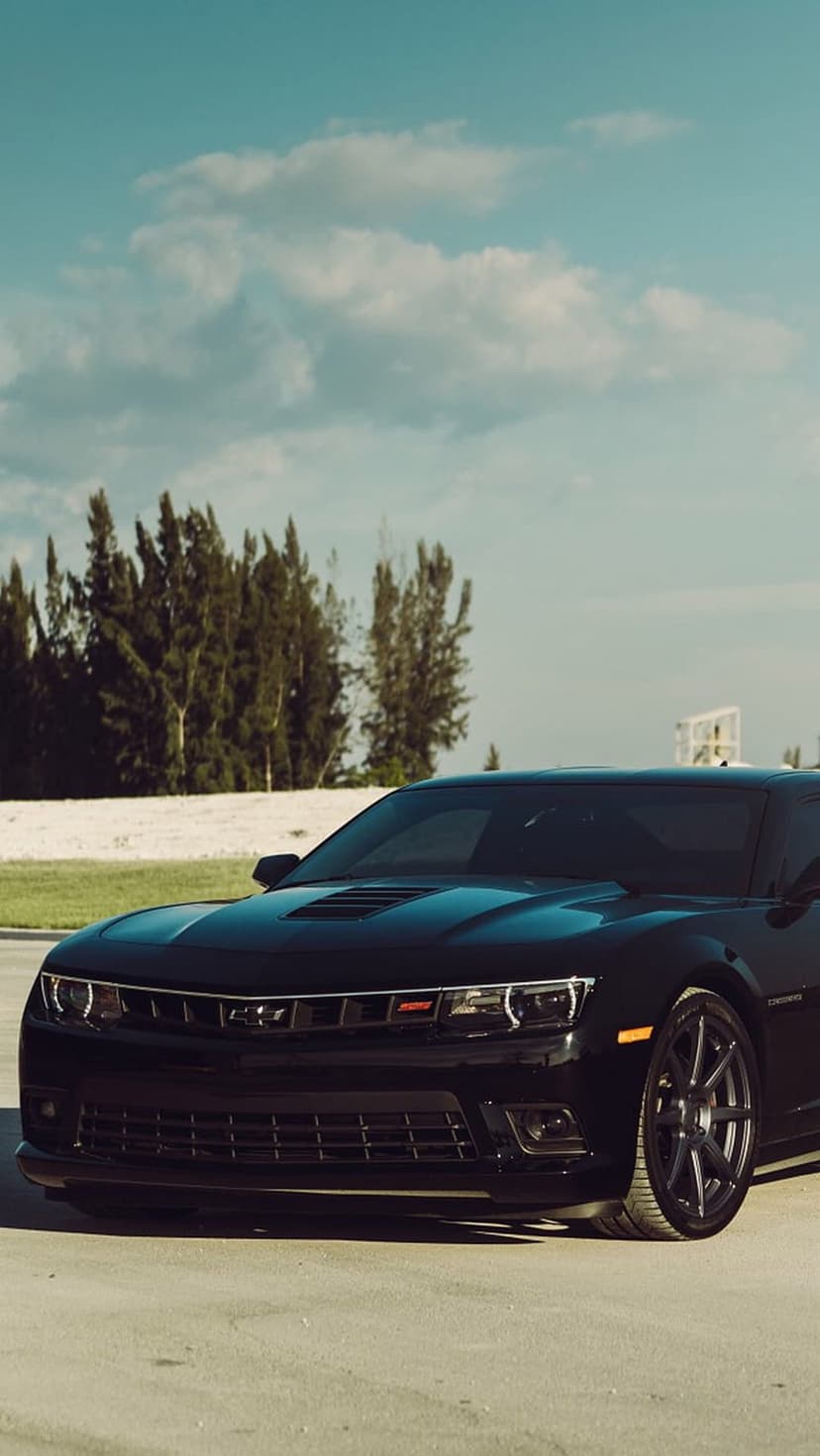 Black Chevrolet Camaro SS with custom wheels parked on a sandy coastal area under a heavy overcast sky.