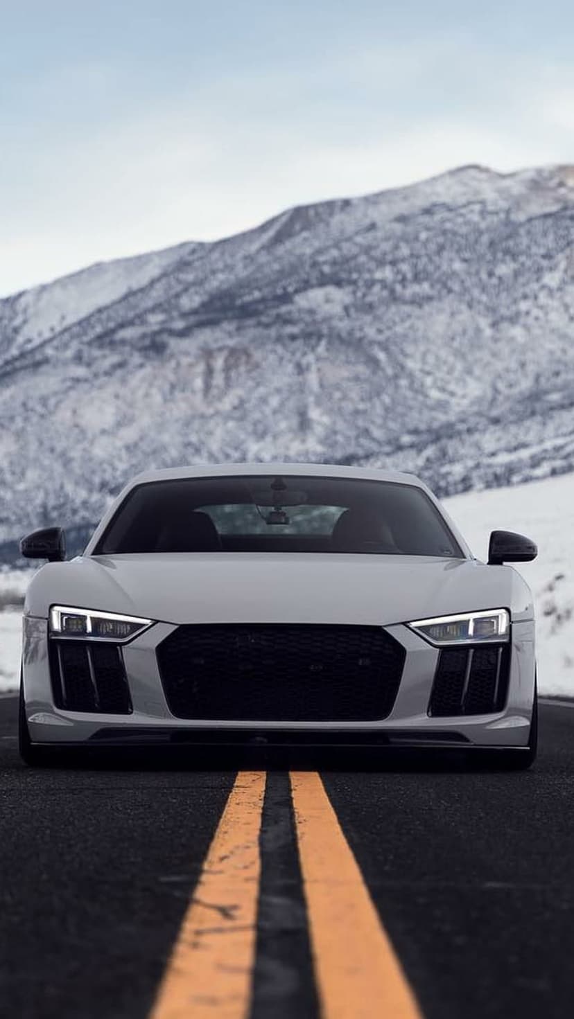 White Audi R8 on an asphalt road with yellow lines, set against a backdrop of snowy mountains and a hazy sky.