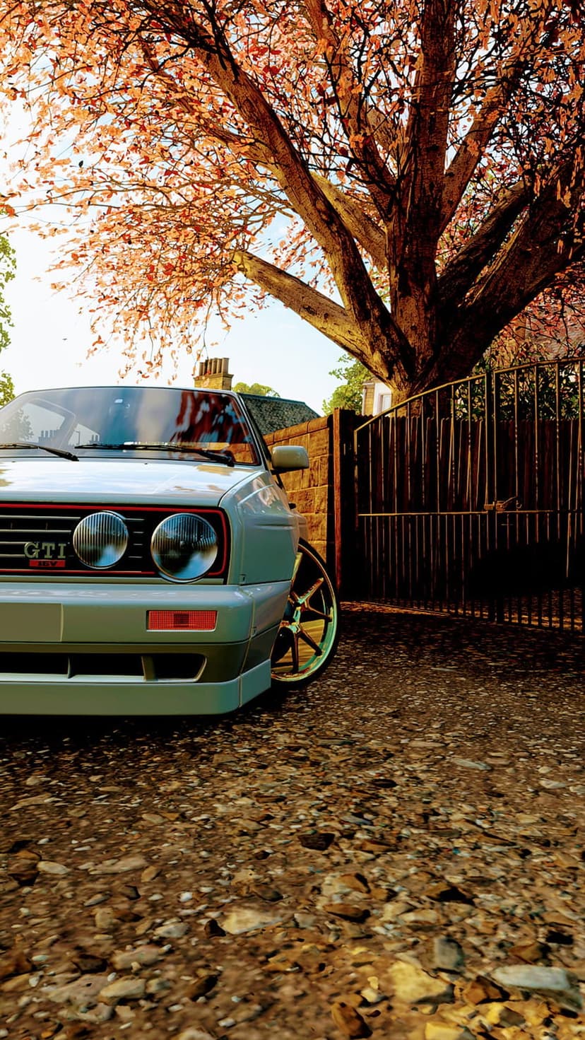 Silver Volkswagen Golf GTI Mk2 parked on a gravel driveway by a stone wall under amber autumn tree leaves.