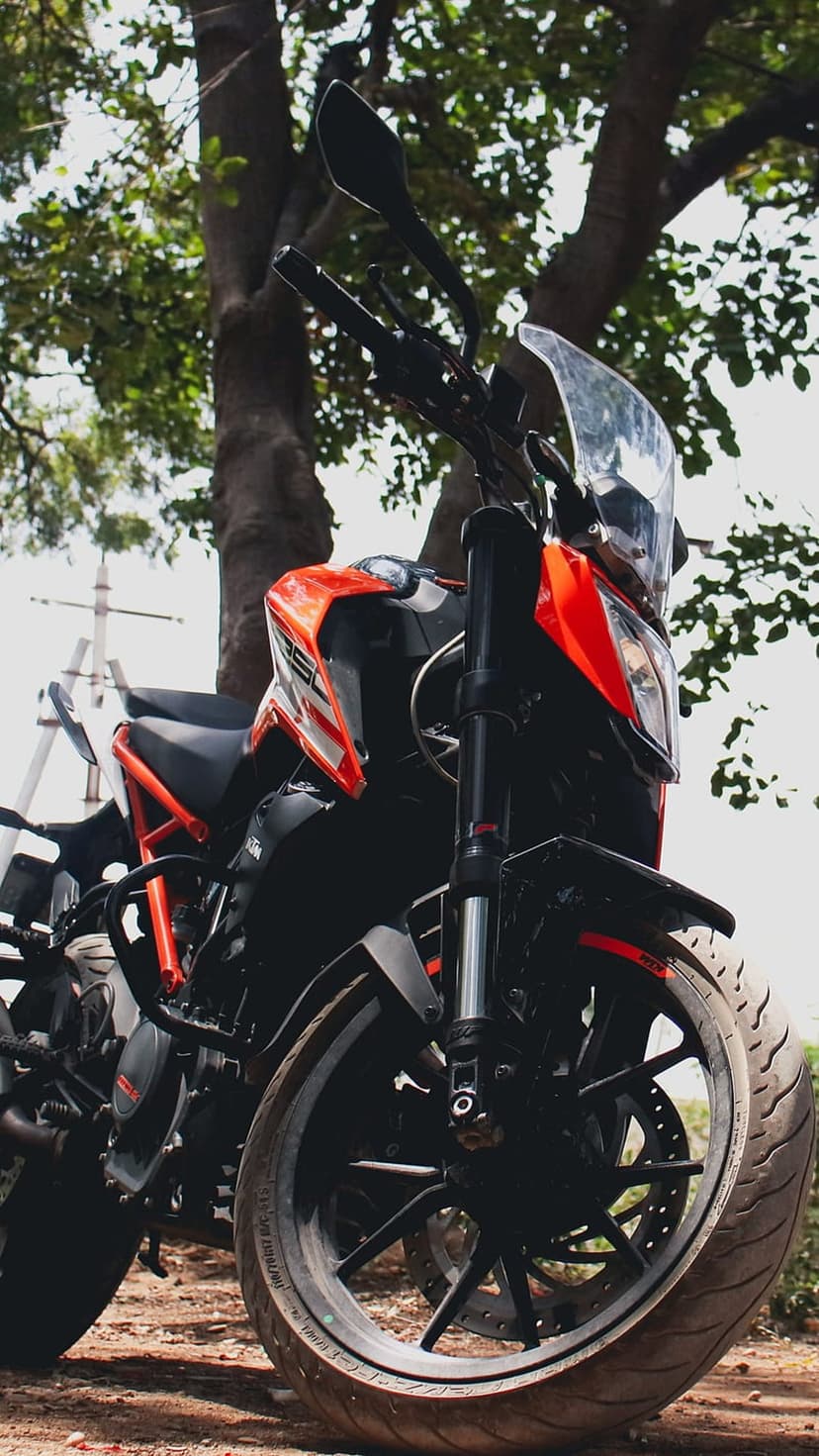 Orange KTM Duke motorcycle with black wheels parked on a dirt trail surrounded by green forest trees.