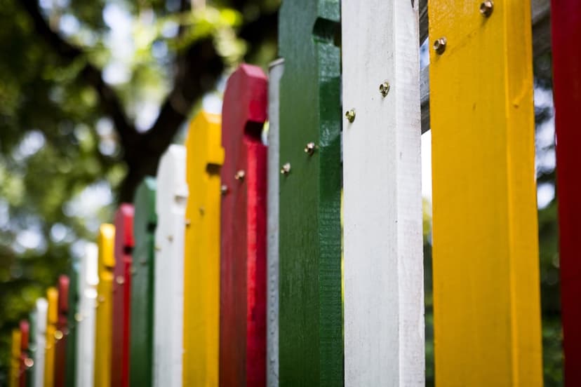 Vertical wooden fence posts in red, green, yellow, and white against a blurred green leafy backdrop.