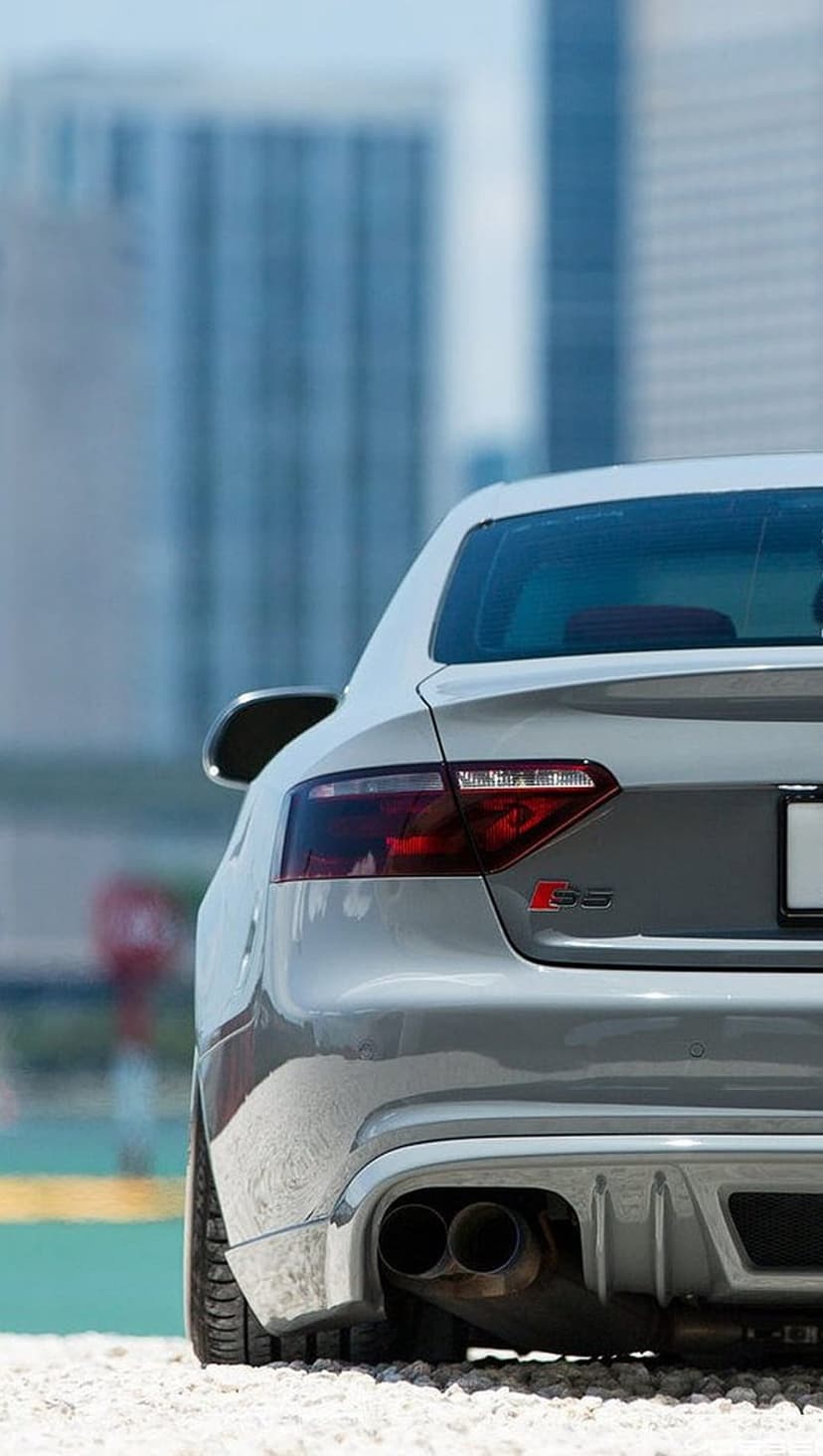 Silver Audi S5 rear bumper featuring red taillights and quad exhausts on gravel with a blurred city background.