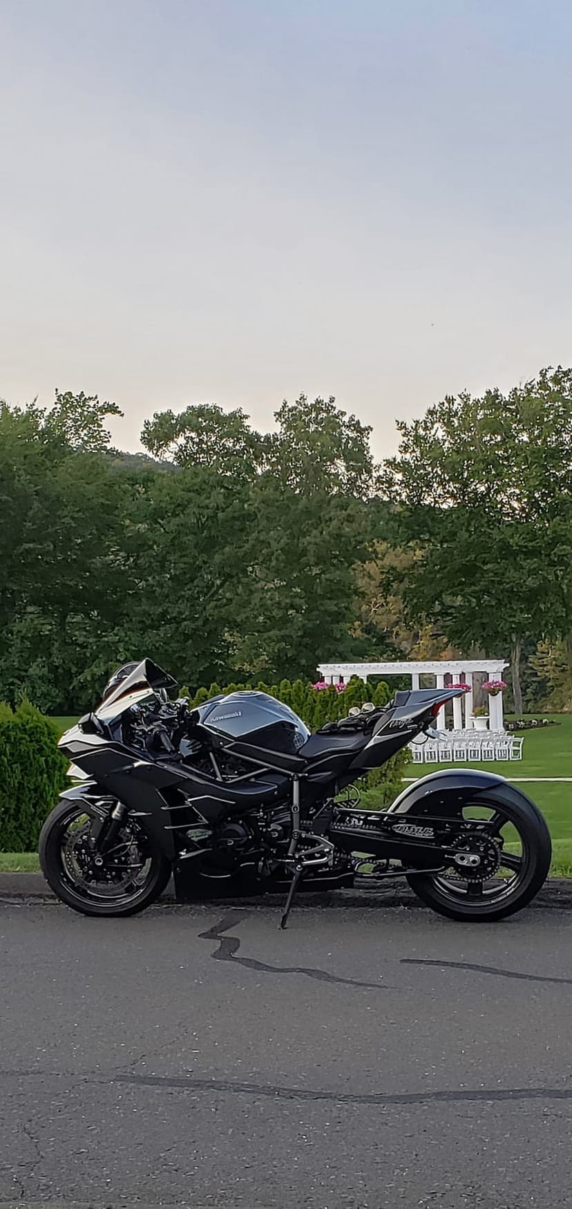 Black Kawasaki motorcycle parked on asphalt with green trees and a white building under soft morning sunlight.