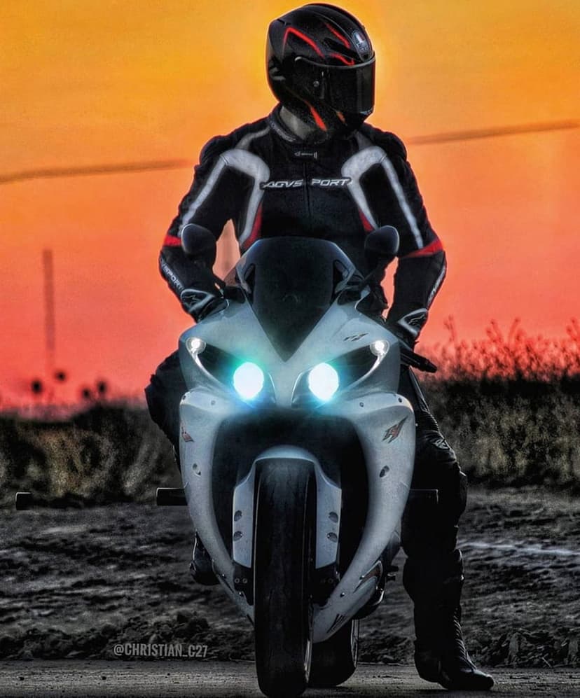 Motorcyclist on a white sportbike with glowing blue headlights against a vibrant orange and red sunset sky.
