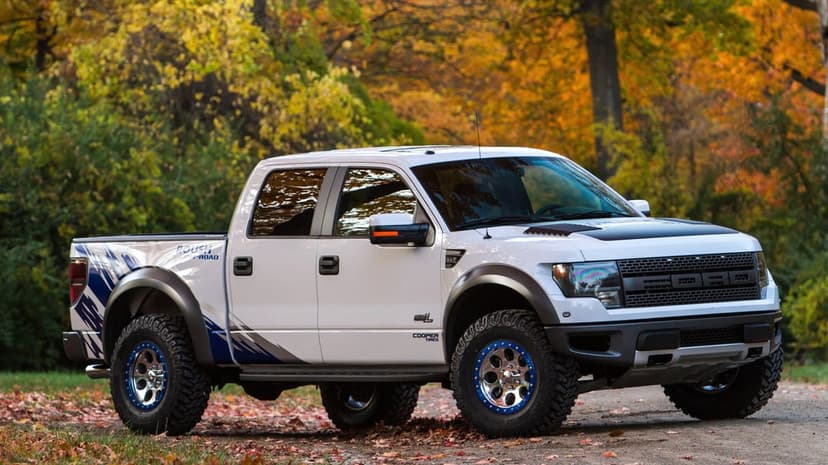 White Ford Raptor truck with blue wheels on a dirt road surrounded by yellow and orange autumn trees.