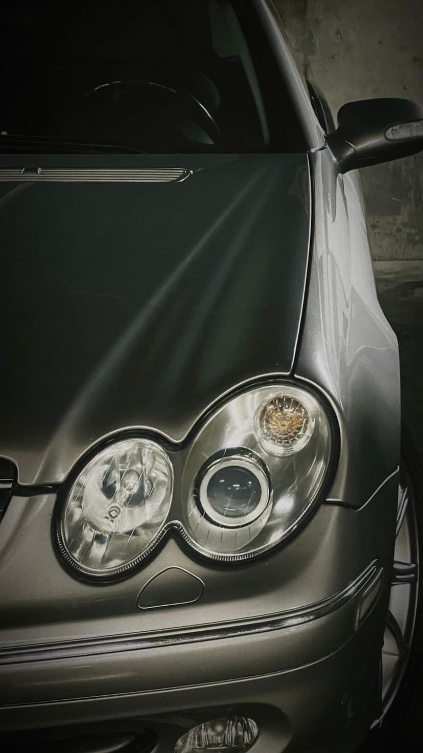 Silver Mercedes-Benz CLK headlight and hood with metallic reflections and projector lens in a low-angle view.