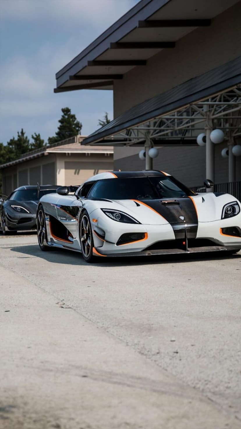 White Koenigsegg hypercar with orange accents parked on asphalt in front of a modern building with a covered walkway.