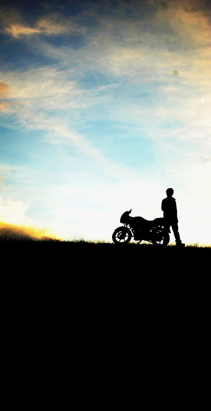 Biker silhouette stands beside a motorcycle on a hill under a crescent moon and colorful sunset sky.
