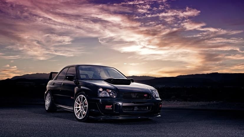 Black Subaru WRX STI parked on asphalt with a dramatic purple sunset and mountain silhouette in the background.