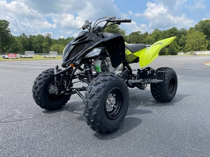 Black and neon yellow Yamaha Raptor 700R ATV parked on a sunny asphalt lot with trees under a blue cloudy sky.
