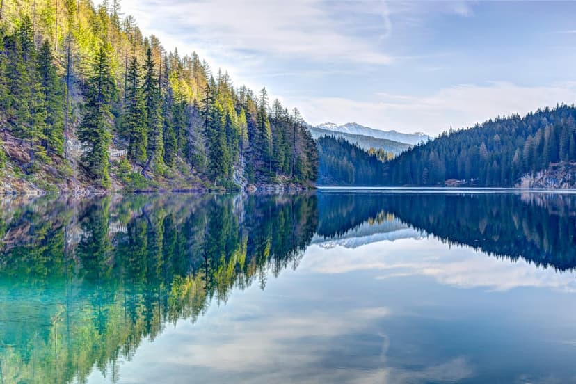 Towering pine trees and jagged mountains reflect in a still alpine lake under a bright blue, wispy sky.