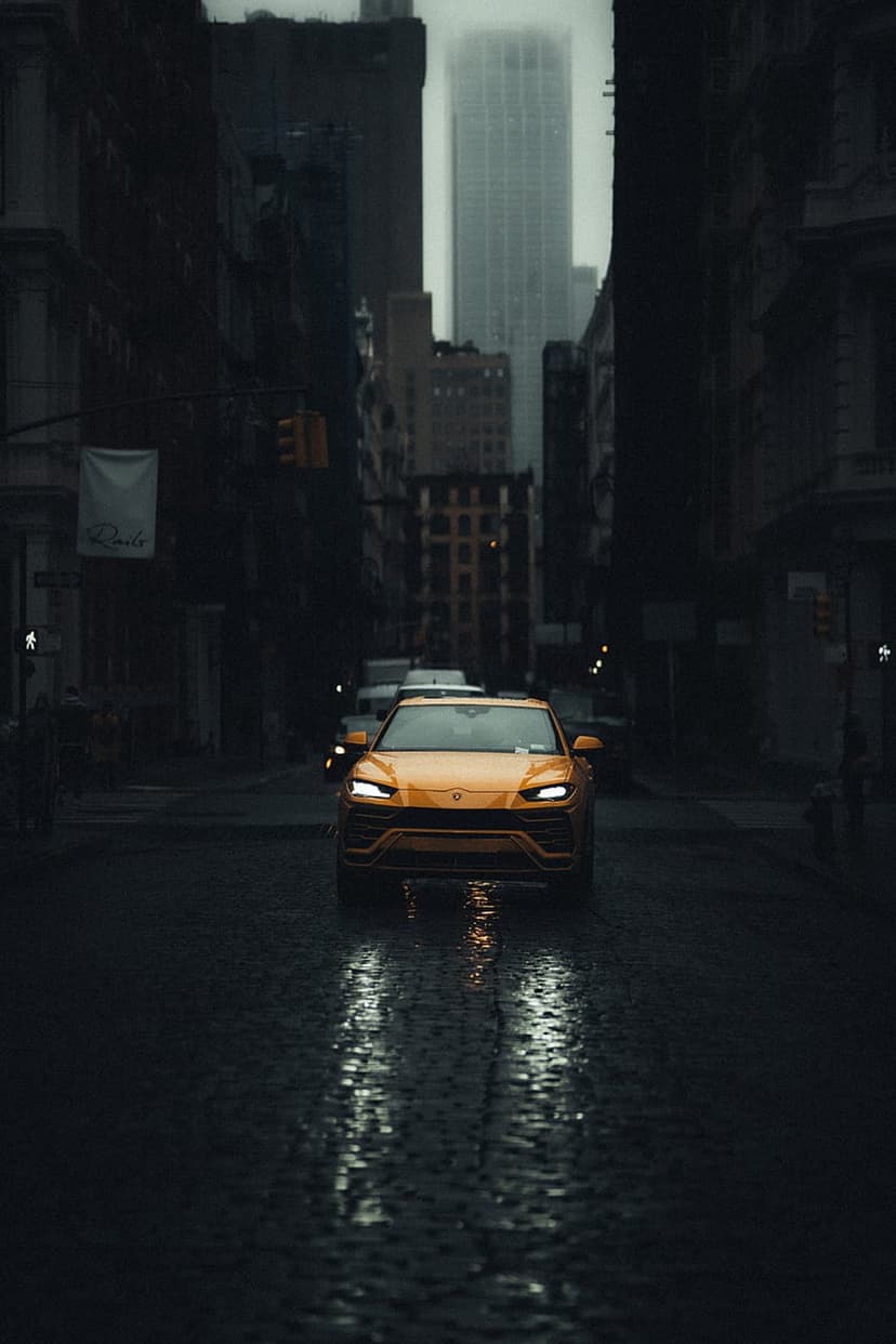 Yellow Lamborghini Urus driving on a wet cobblestone street at dusk with glowing headlight reflections.