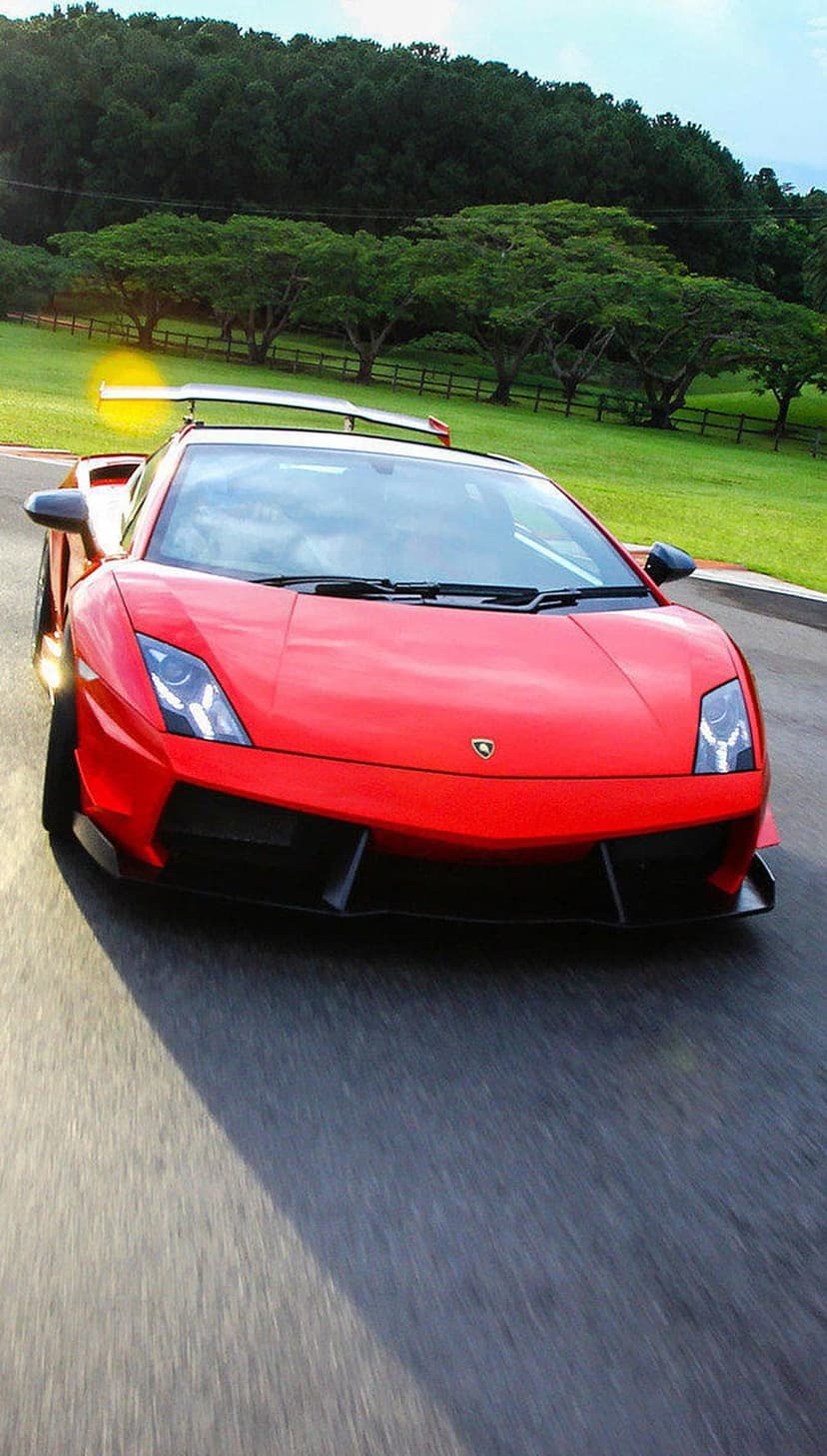 Red Lamborghini Gallardo speeds along a grey racetrack surrounded by lush green trees and rolling grassy hills.