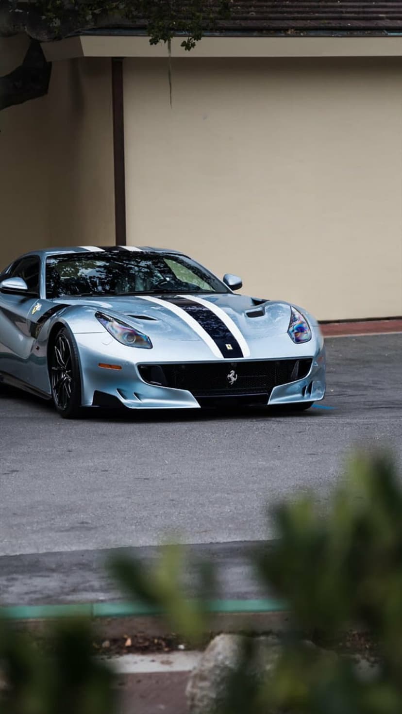Silver Ferrari F12 Berlinetta with black and white racing stripes parked on asphalt near a neutral building.