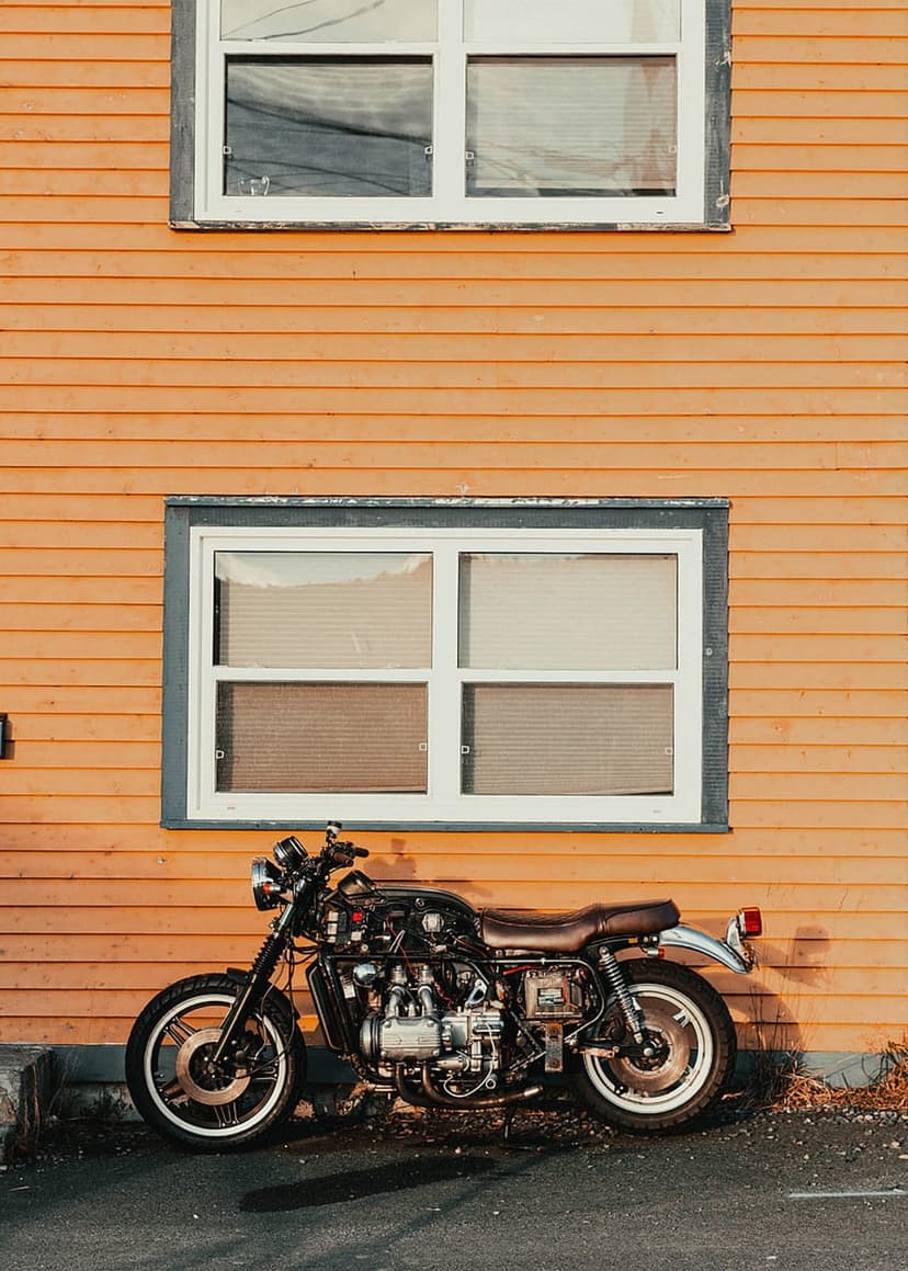 Vintage black motorcycle with a brown leather seat parked against bright orange house siding with white windows.