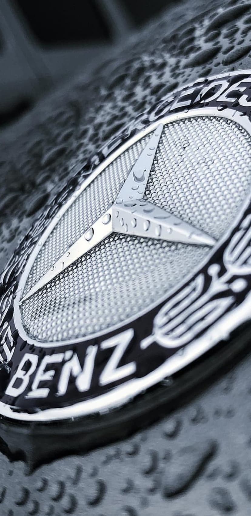 Mercedes-Benz three-pointed star logo covered in clear water droplets against a dark textured car surface.