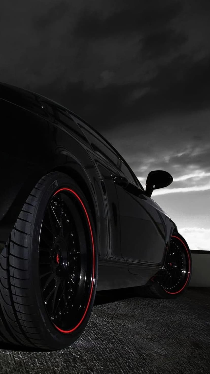 Black supercar wheel with a red rim accent sits under a dark, dramatic sky with moody cloud formations.