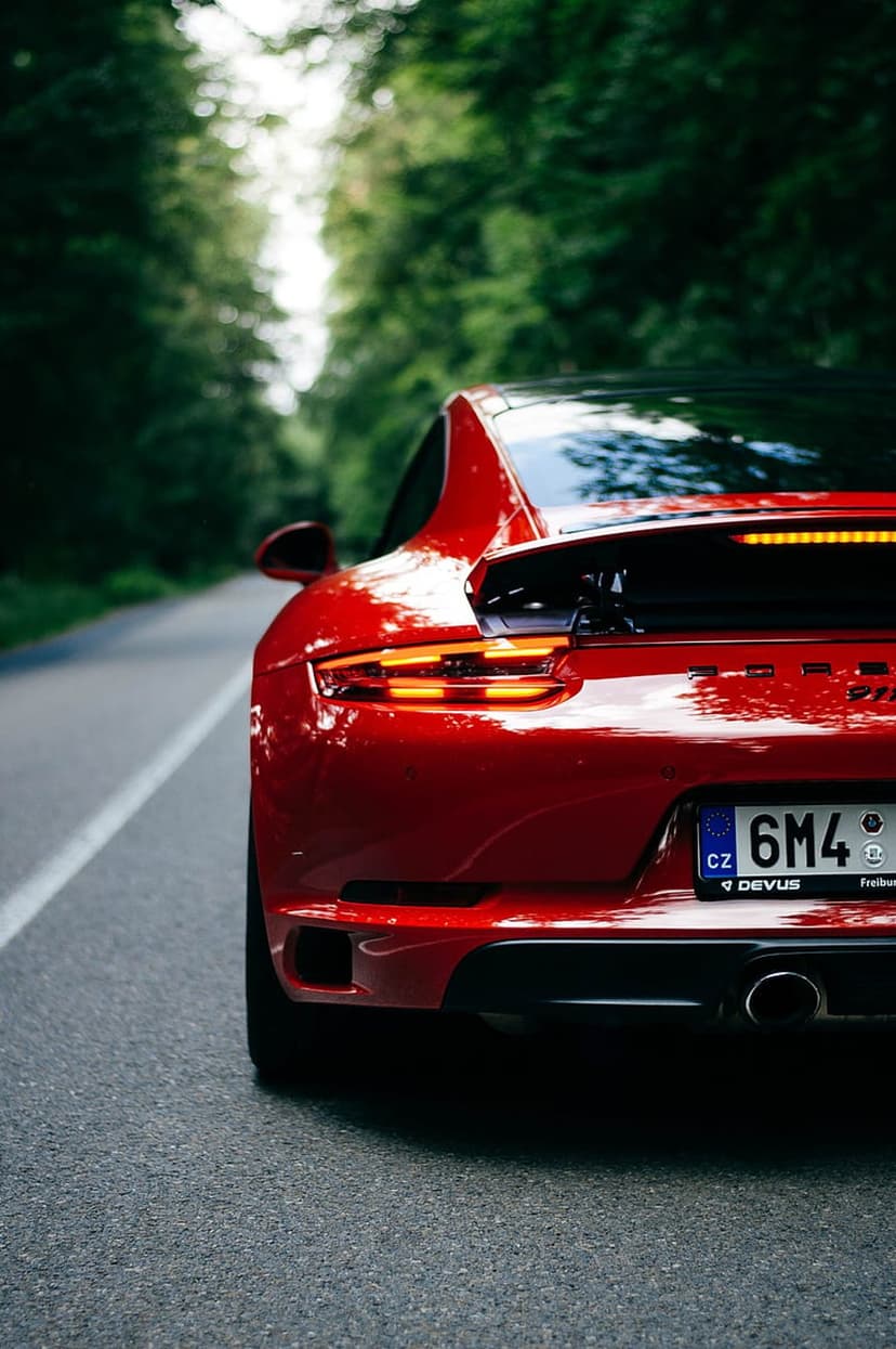 Red Porsche rear view with glowing taillights driving on a paved road through a dense green forest landscape.