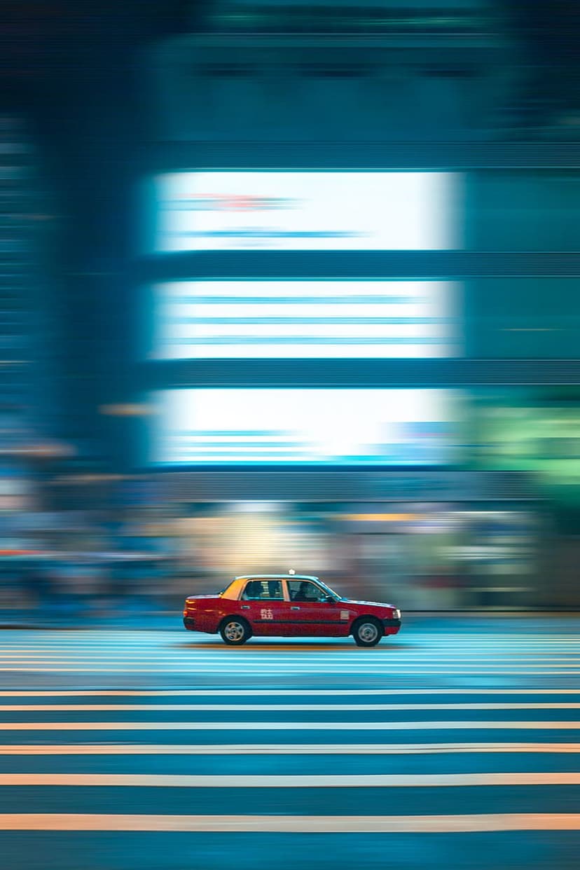 Red taxi cab speeds through a city crosswalk at night with vibrant neon light trails and blurred skyscrapers.