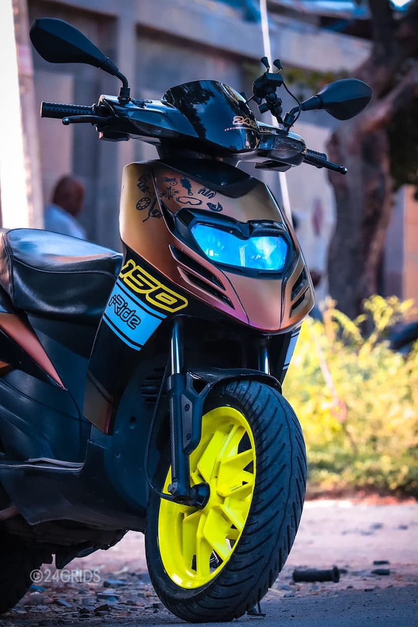 Modern scooter front with glowing blue LED headlights, bronze bodywork, and bright yellow rims in close-up view.