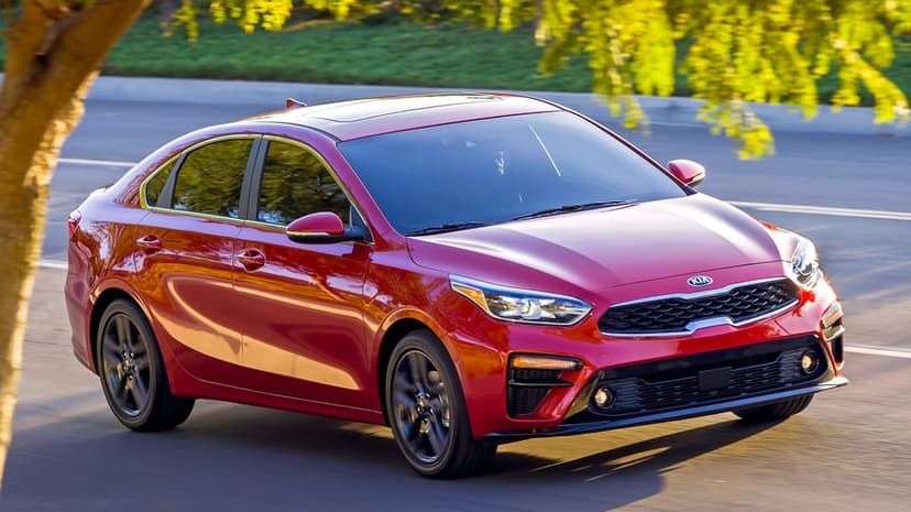Red Kia Forte sedan driving on a sunlit asphalt road with blurred green and yellow trees in the background.