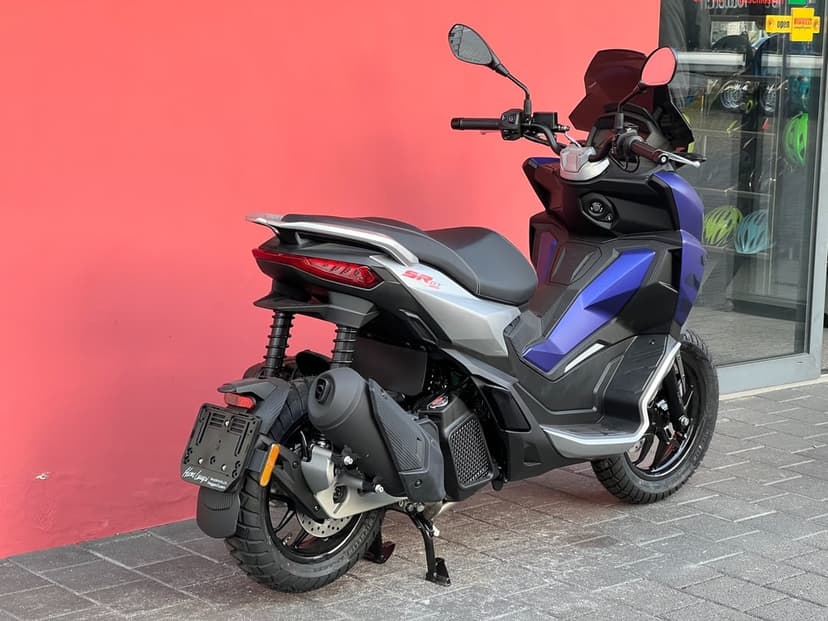 Rear view of a matte blue and silver Aprilia SR GT scooter parked on pavement against a bright red wall.
