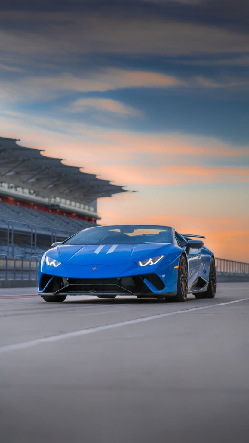 Blue Lamborghini convertible on a race track under a sunset sky with a stadium grandstand in the background.