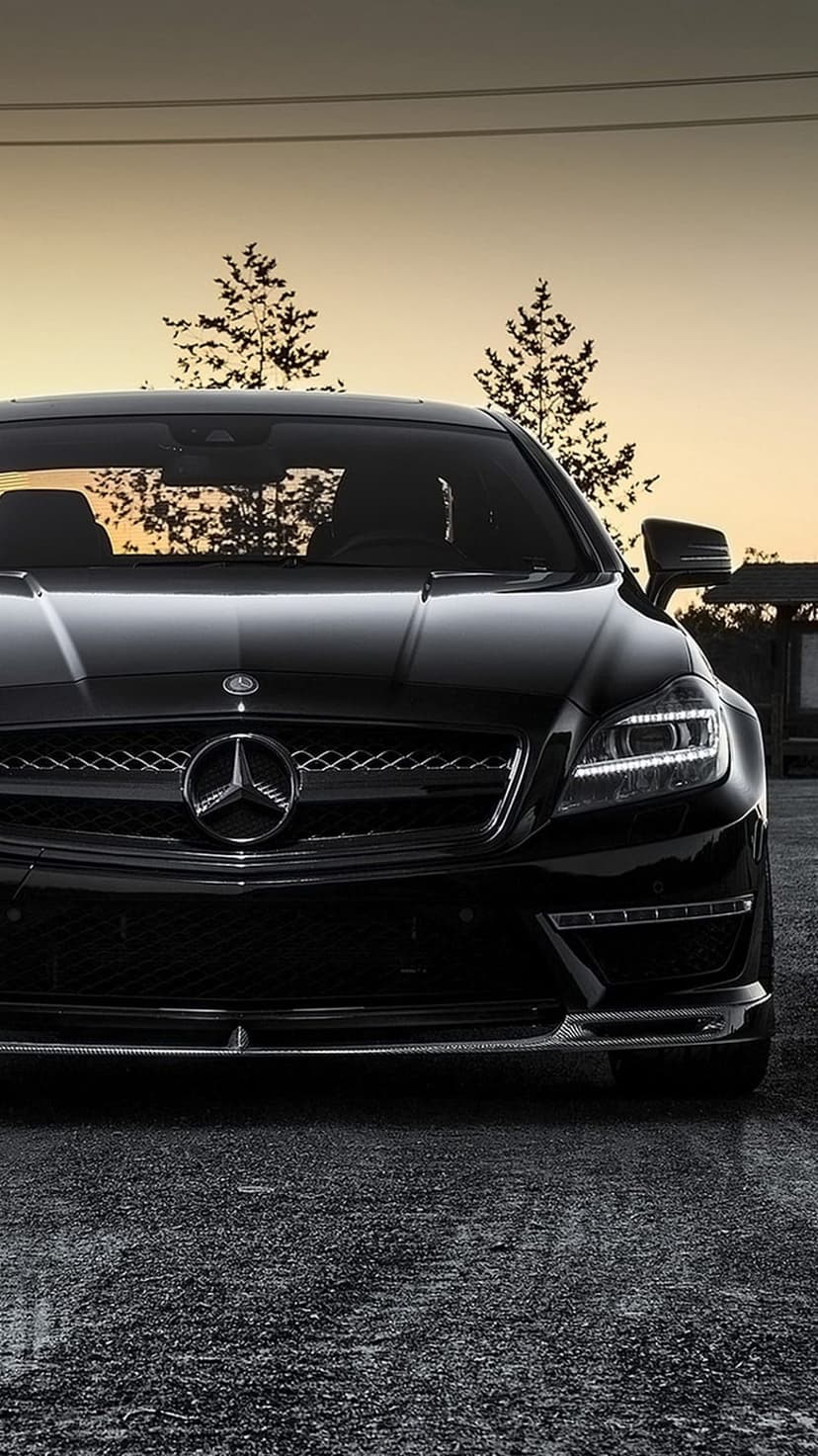 Mercedes-Benz CLS coupe parked on wet asphalt during golden hour with glowing LED headlights and a sunset background.
