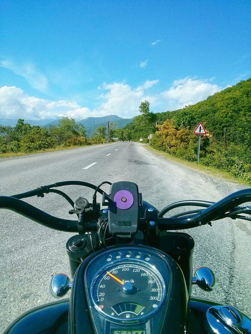 Motorcycle rider's view of a winding road through green hills and mountains under a bright blue sky.