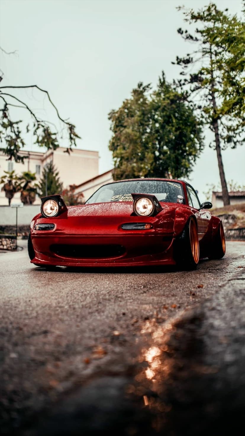 Red Mazda Miata with pop-up headlights on a wet road, widebody kit, and bronze wheels under overcast light.