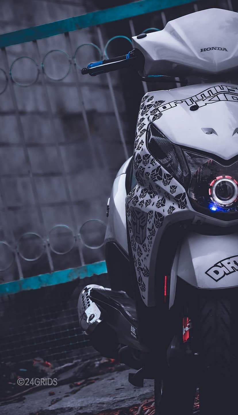 White Honda scooter with a red Autobot decal and bright LED headlight against a blurred teal gate background.