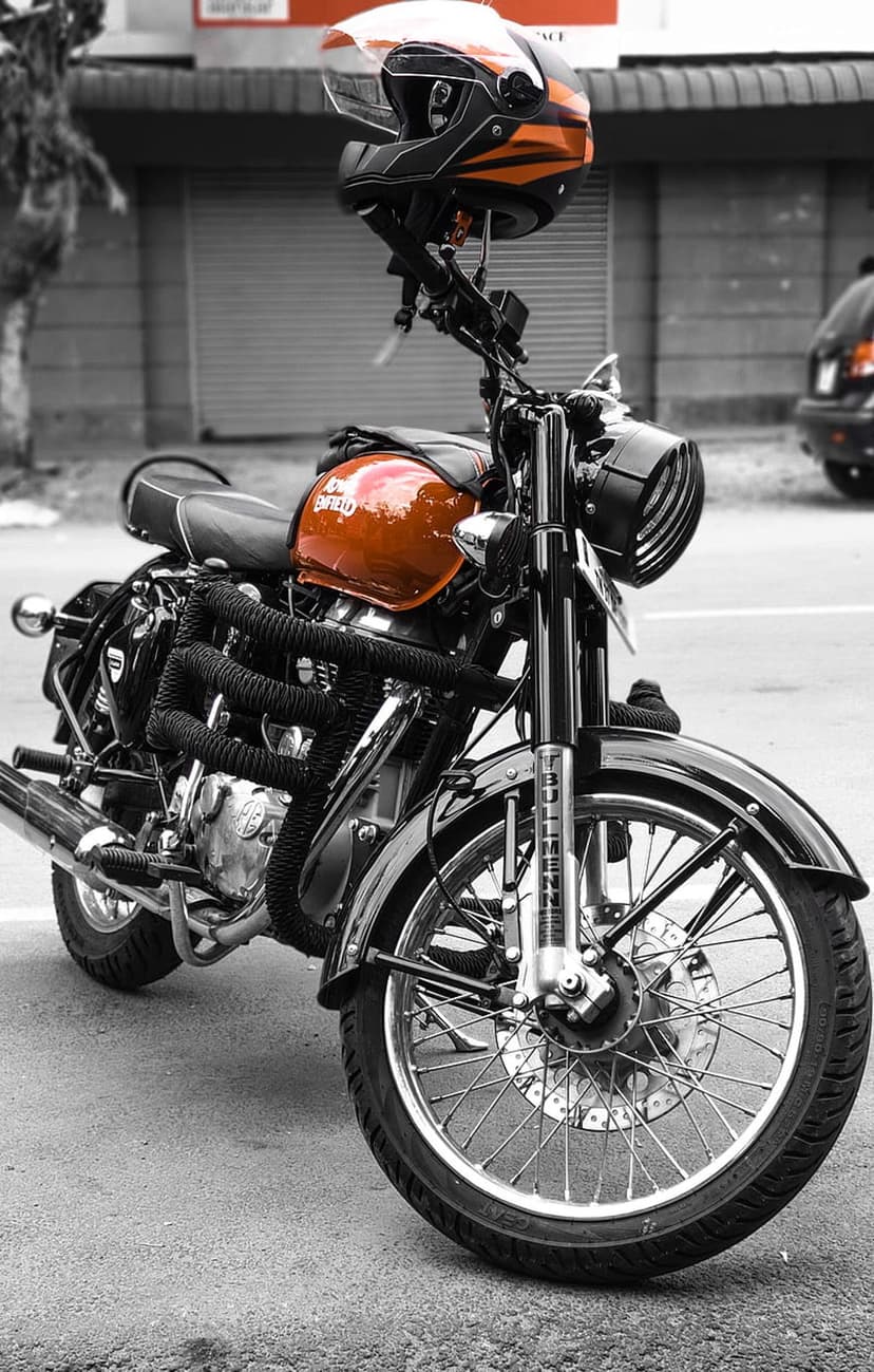 Orange Royal Enfield motorcycle with a matching helmet on the handlebars against a blurred grayscale street.