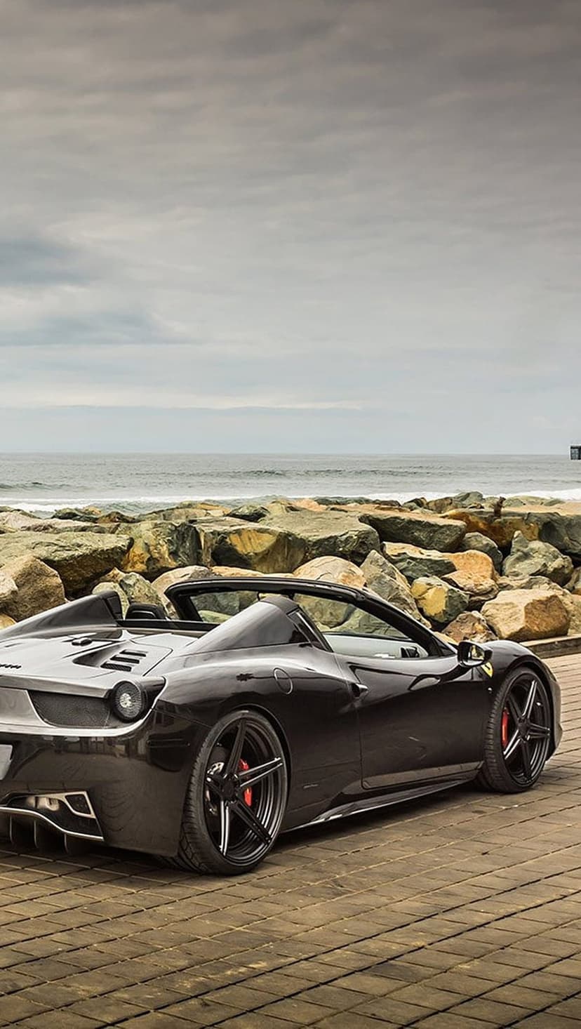 Black Ferrari convertible parked on a wooden deck overlooking a rocky coastline and a misty grey ocean horizon.