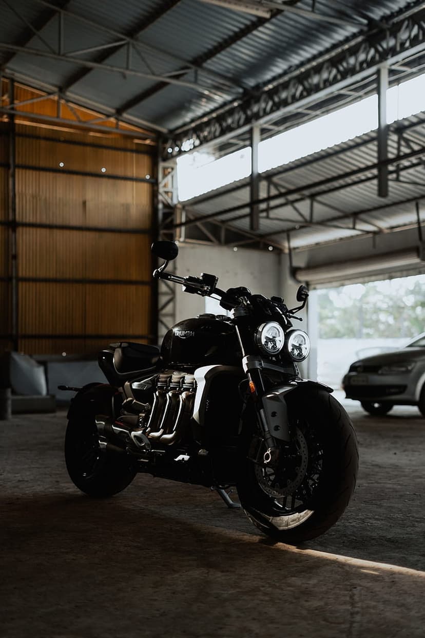 Black Triumph Rocket 3 motorcycle parked in a dark garage featuring chrome exhaust pipes and engine details.