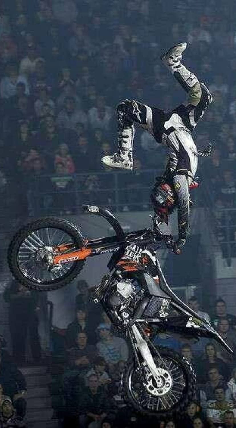 Motocross rider performs a vertical leg-up stunt mid-air while silhouetted against a blurred stadium crowd.