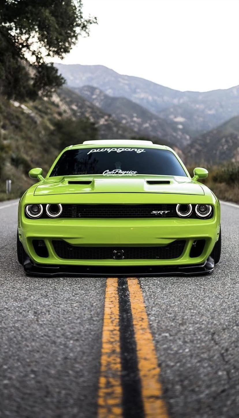 Lime green Dodge Challenger SRT with halo headlights on a winding mountain road under a cloudy, scenic sky.
