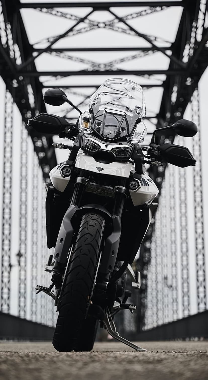 Triumph Tiger adventure motorcycle parked on a concrete road between the metal trusses of a large steel bridge.