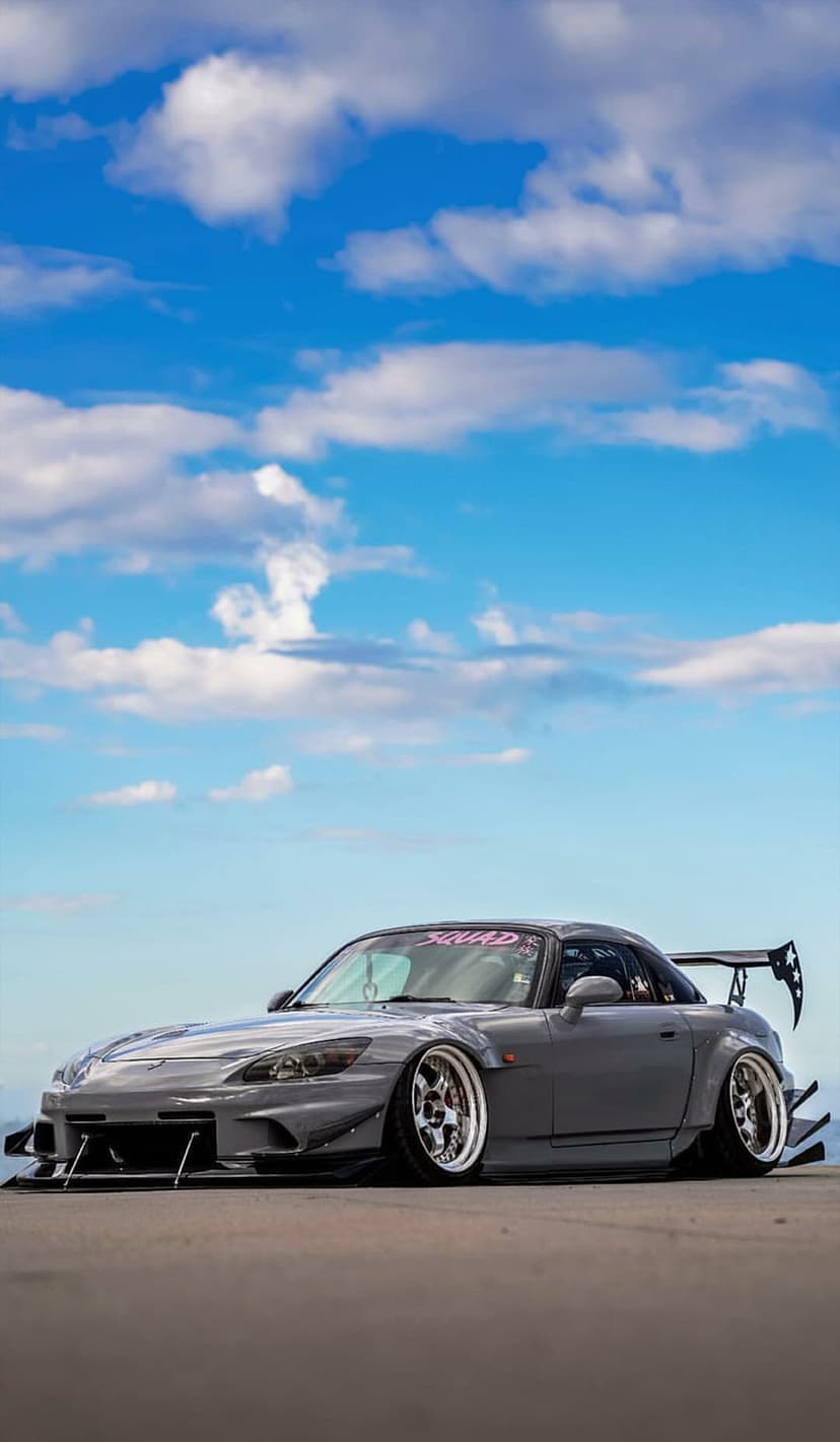 Gray Honda S2000 with a widebody kit and aftermarket wheels parked outdoors under a blue sky with white clouds.