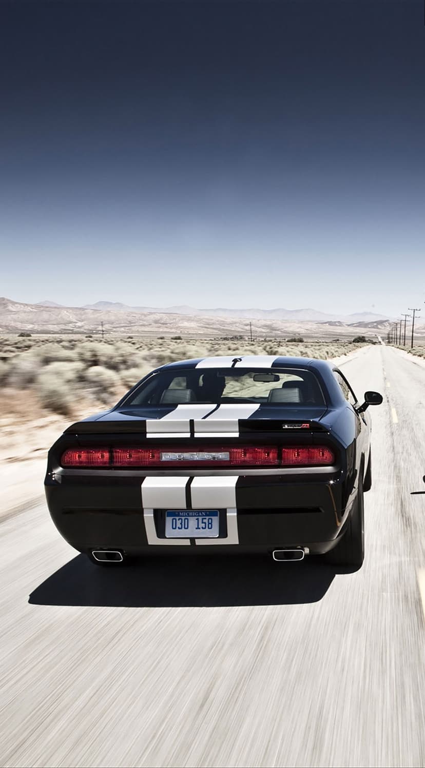 Black Dodge Challenger with white stripes driving on a straight desert highway under a clear blue sky.