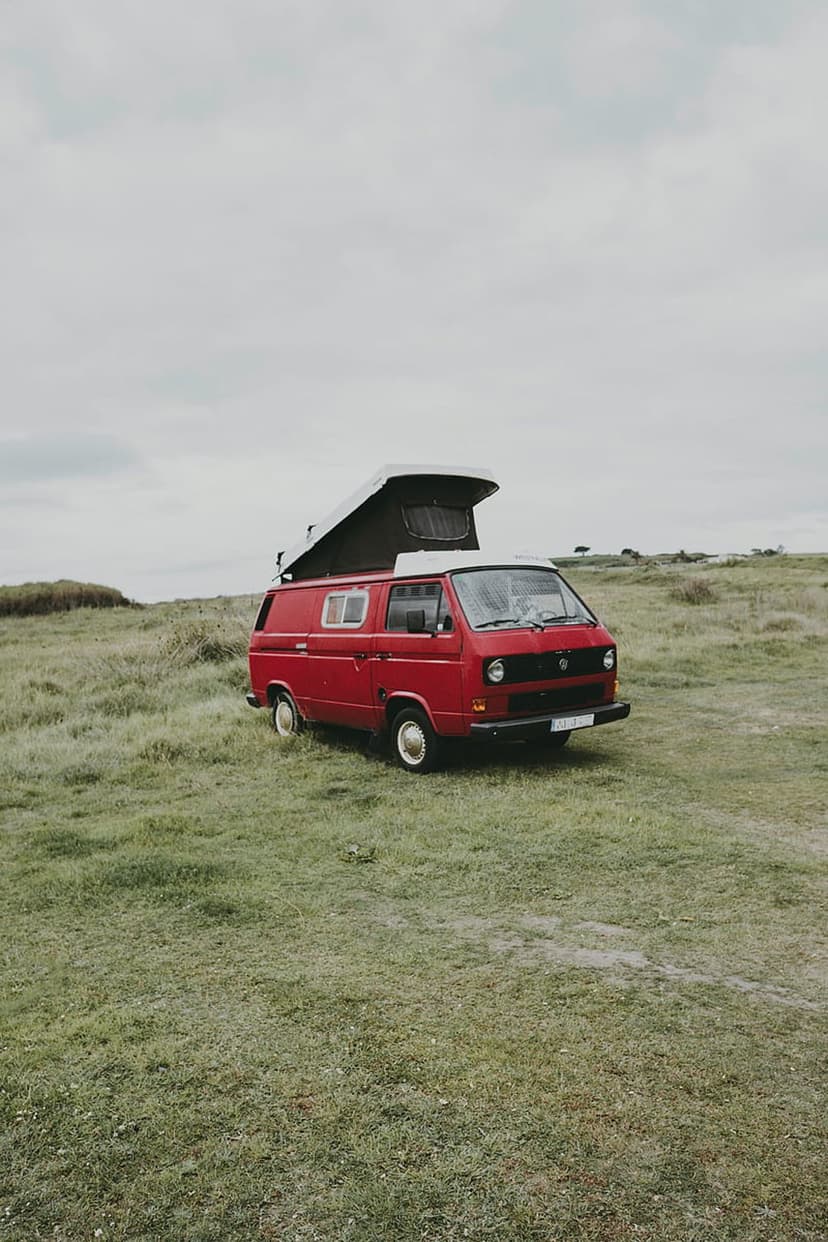 Red vintage camper van with a pop-top roof parked in a vast green field under a cloudy, overcast sky.