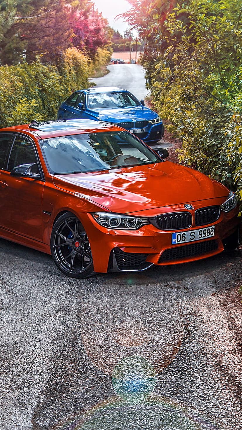Orange BMW sedan leading a blue BMW on a tree-lined asphalt road with sunlight reflecting off the car bodies.