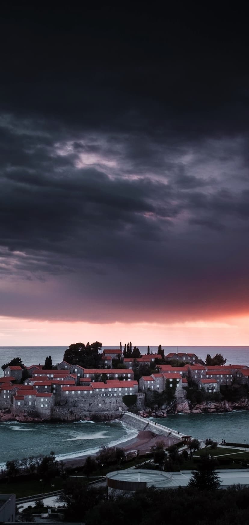 Mediterranean island village with red roofs and a stone causeway under a stormy purple and orange twilight sky.